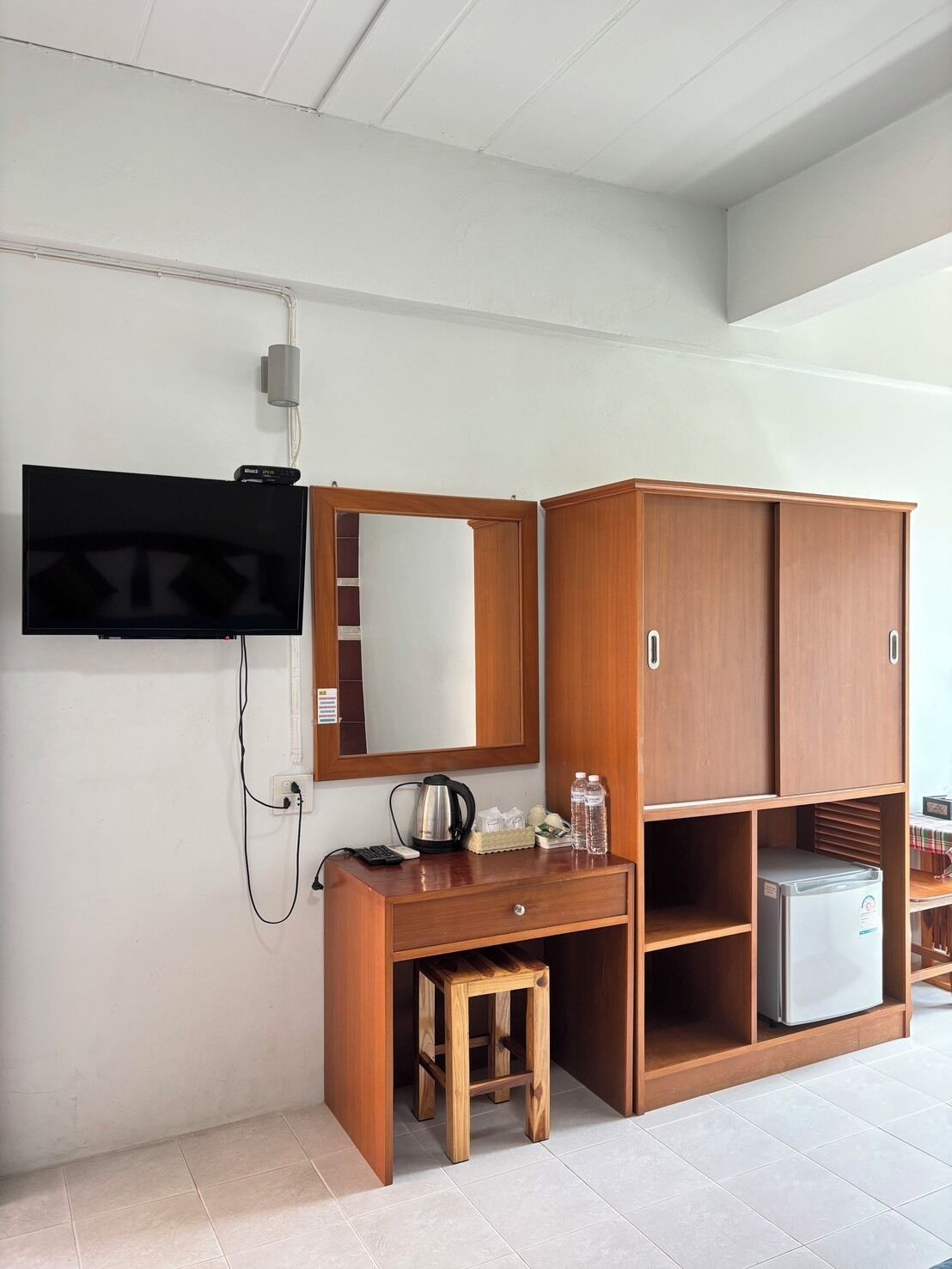 Una camera d'albergo con una scrivania in legno, una televisione a muro, uno specchio, uno sgabello e un mini-frigorifero.