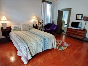 A bedroom with a bed, two bedside lamps, a purple chair, a TV, and a dresser. - Historic Double Shotgun Home in Spanish Town, Downtown Baton Rouge (Baton Rouge)