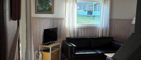 A room with a wooden ceiling, a black leather sofa, a television on a wooden stand, and a window with curtains.
