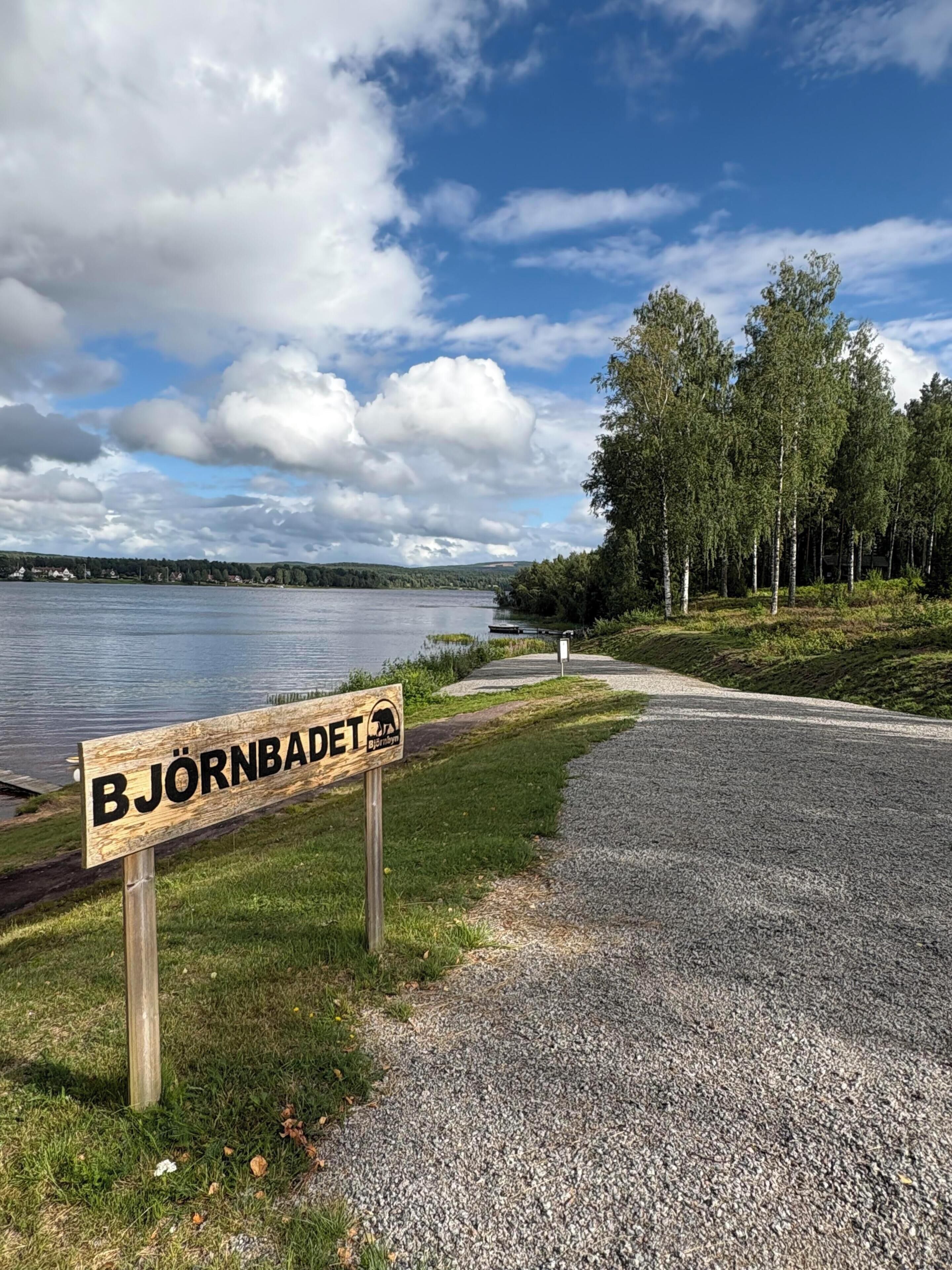 Placa de Björnbadet, uma área natural com trilha de cascalho, árvores e um lago.