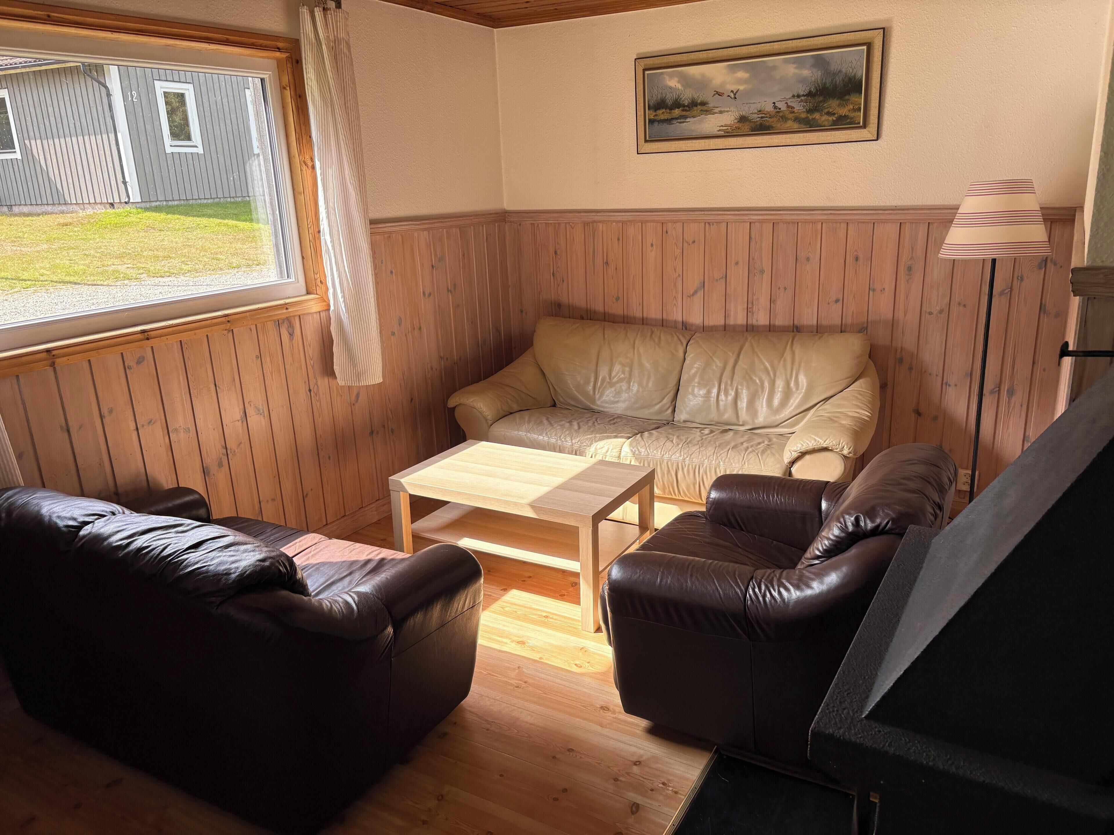 A cozy room with a wooden paneled wall, a beige sofa, a brown leather couch, a wooden coffee table, and a framed landscape painting.
