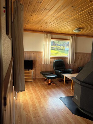 A cozy room with wooden flooring, a television, a leather chair, a small table, and a window with curtains. - Björnbyn Stugby (Värmlands län)