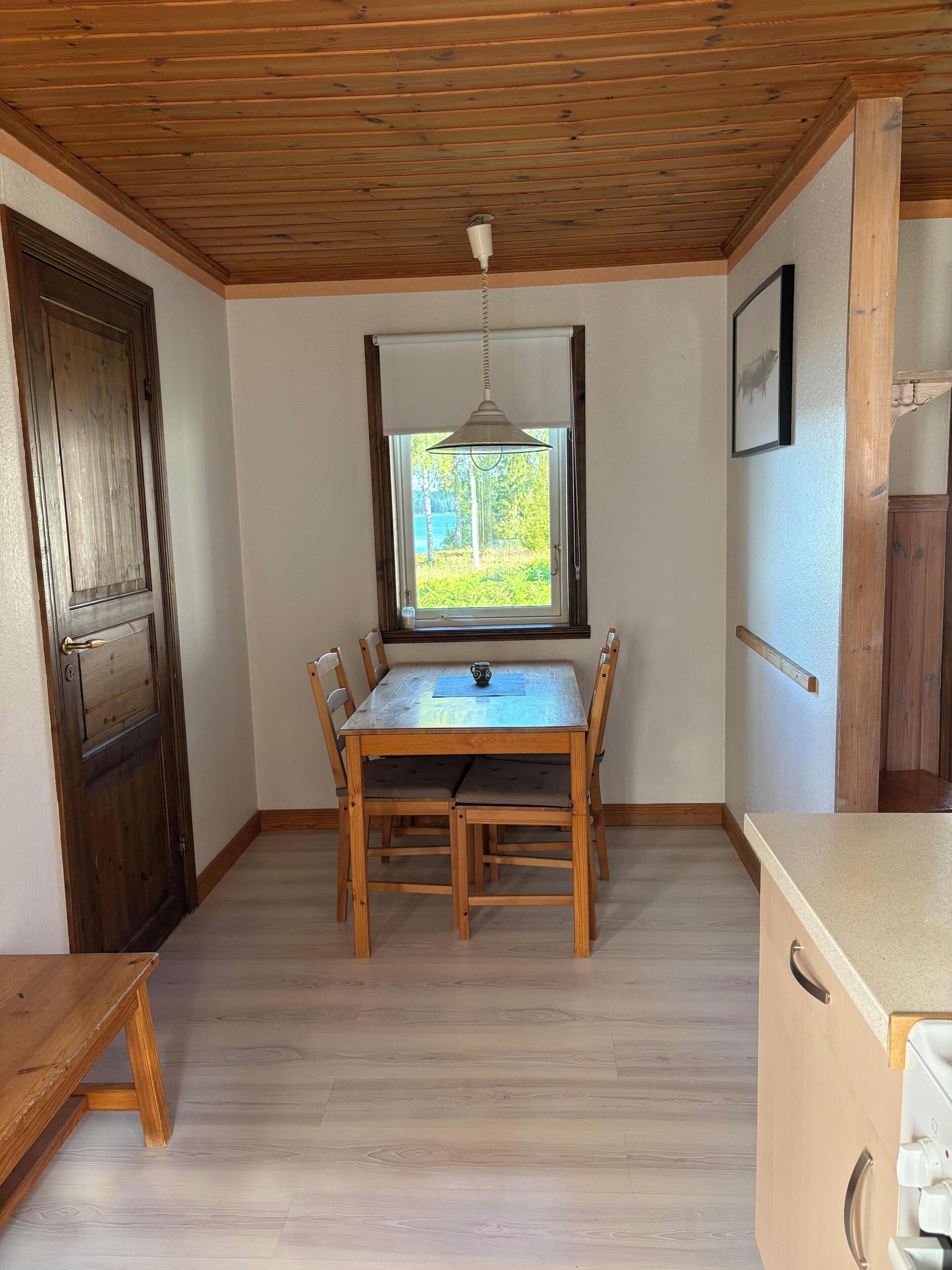 A room with a wooden ceiling, a dining table with chairs, a window with a view of greenery, and a door.
