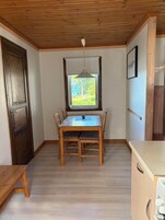 A room with a wooden ceiling, a dining table with chairs, a window with a view of greenery, and a door.