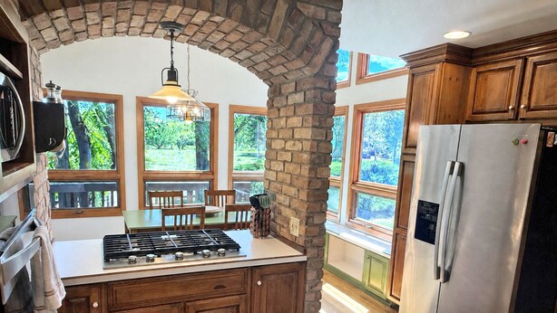 A kitchen with a central island, wooden cabinets, a stainless steel refrigerator, and large windows with a view of greenery.