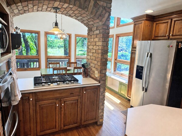 A kitchen with a central island, wooden cabinets, a stainless steel refrigerator, and large windows with a view of greenery.