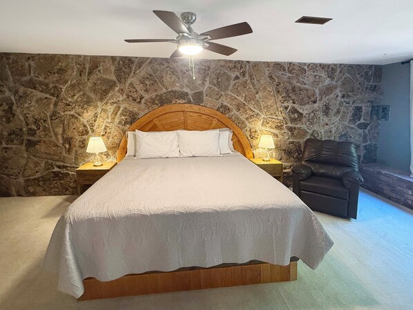 A bedroom with a stone wall, a wooden bed frame, two bedside lamps, and a ceiling fan.