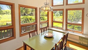 A dining area with a table, chairs, and a chandelier. There are multiple windows with views of greenery.