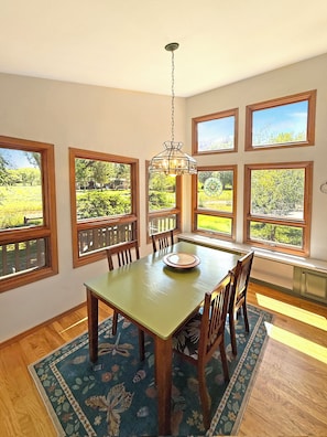 A dining area with a table, chairs, and a chandelier. There are multiple windows with views of greenery.