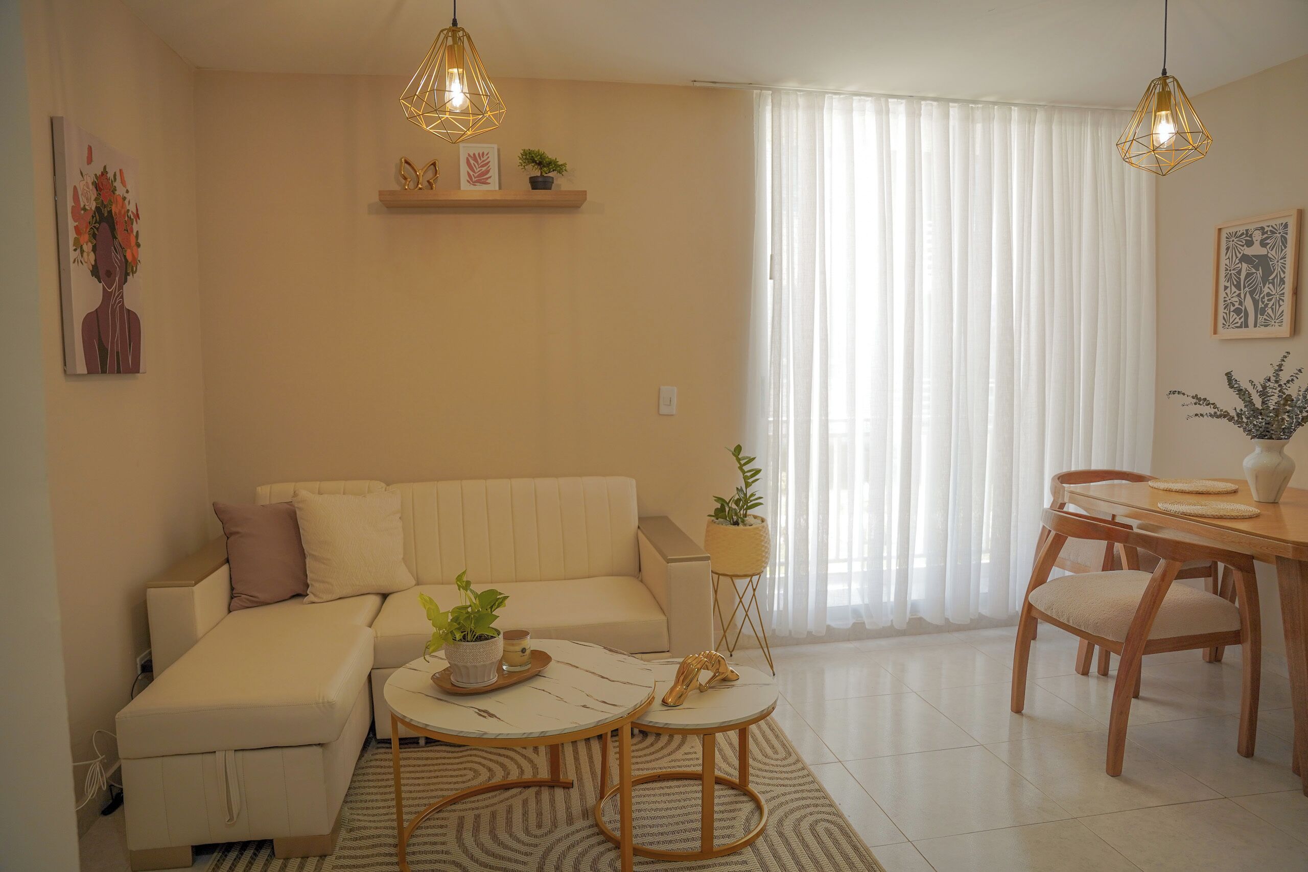 A living room with a sofa, a coffee table, a dining table with chairs, and a large window with sheer curtains.