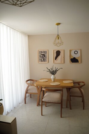 A dining area with a wooden table, four chairs, a vase with dried plants, and three framed artworks on the wall.