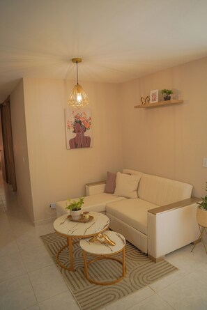 A modern living room with a beige sofa, two round tables with gold accents, a hanging light fixture, and a wall-mounted shelf with decorative items.