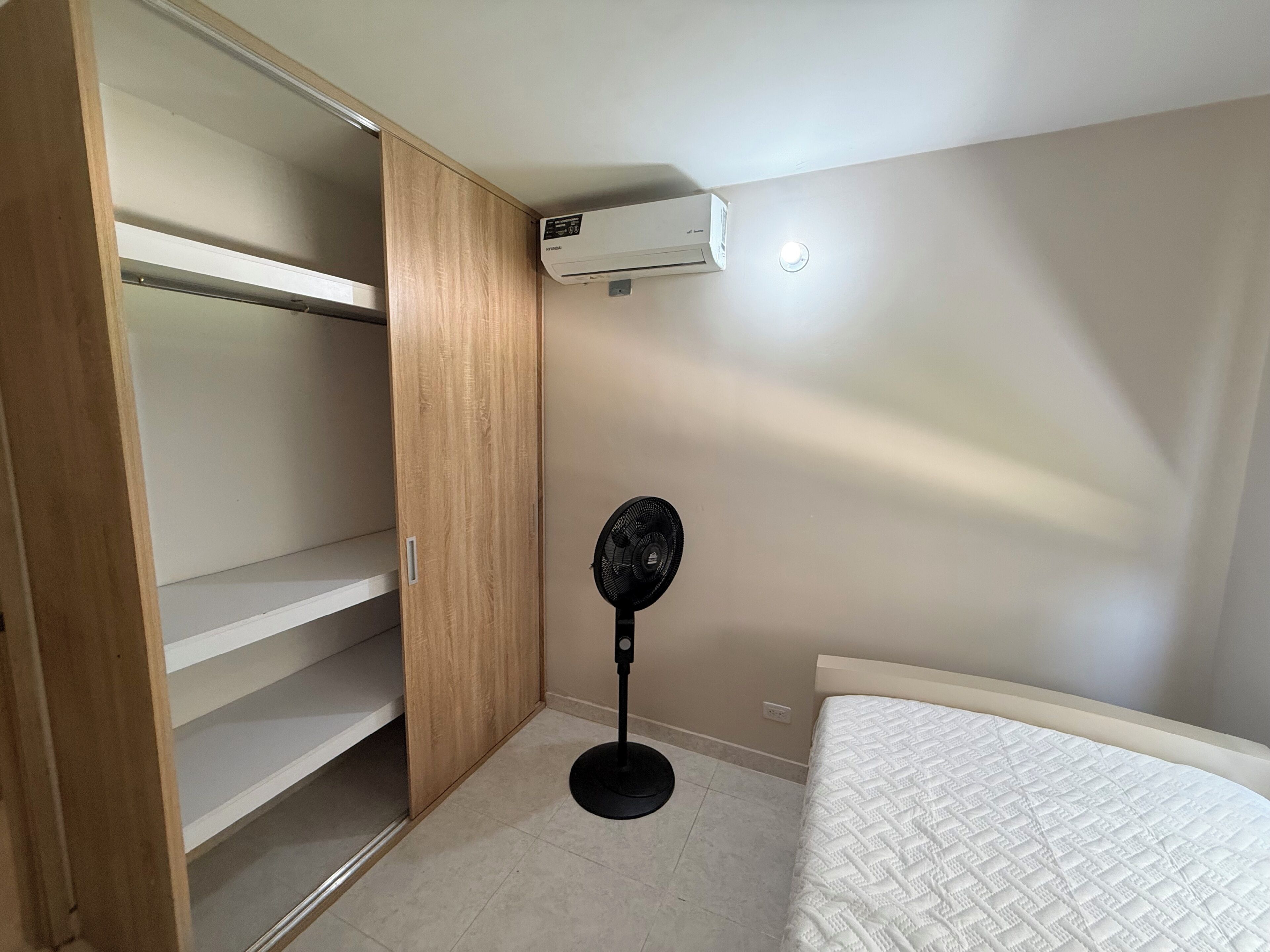 A bedroom with a bed, a standing fan, a wardrobe, and an air conditioning unit.