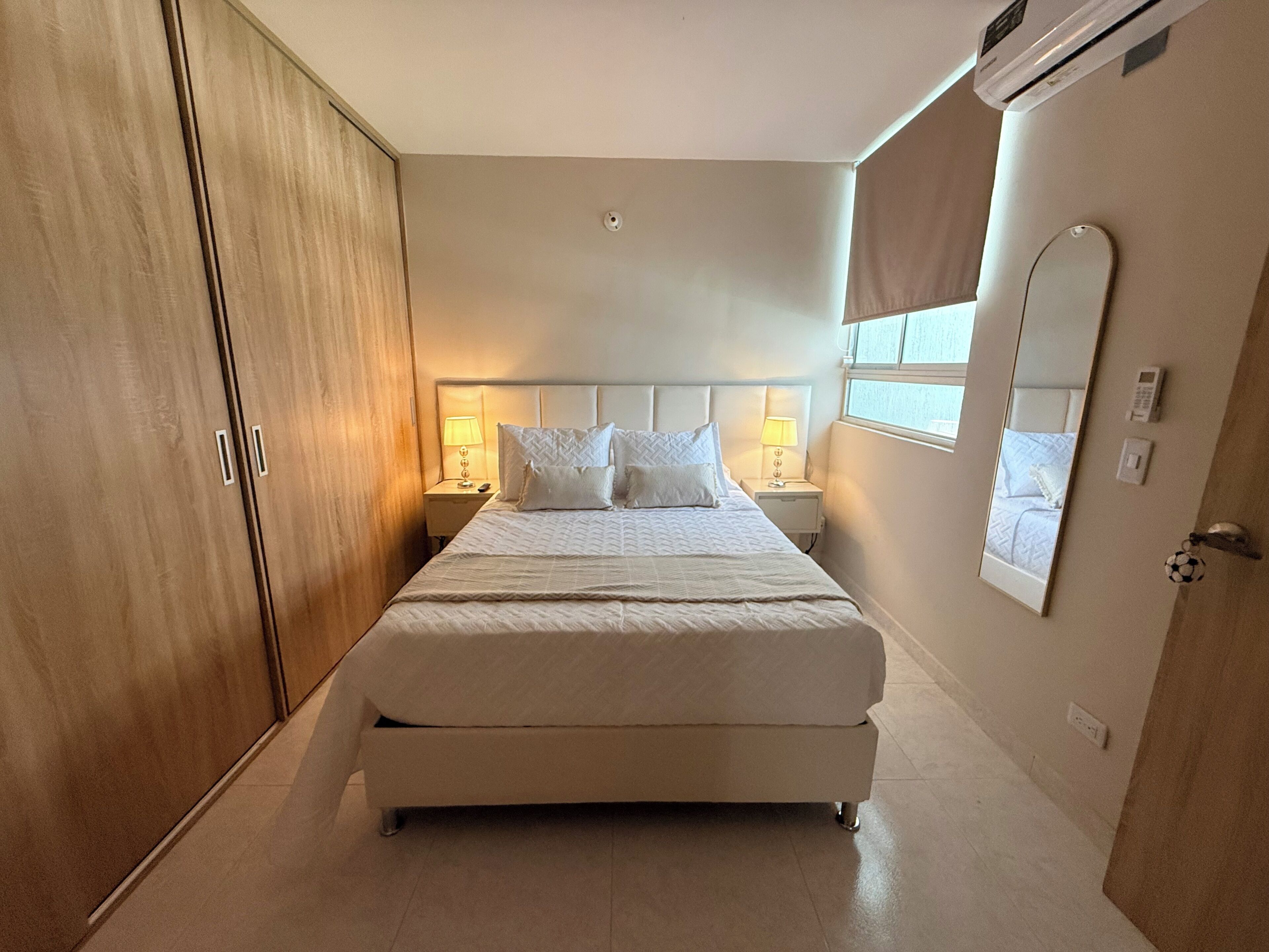 A hotel room with a large bed, bedside lamps, a wardrobe, a mirror, and an air conditioning unit.