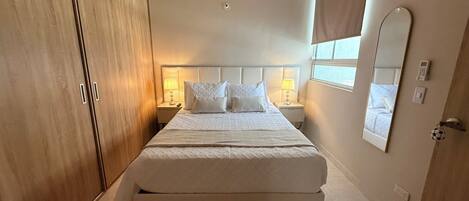 A hotel room with a large bed, bedside lamps, a wardrobe, a mirror, and an air conditioning unit.