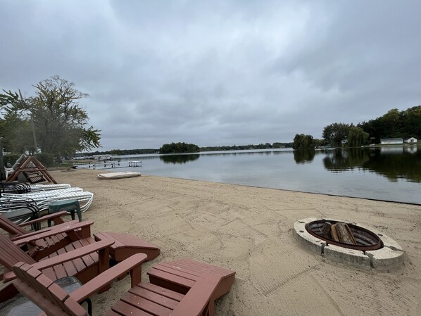 A lakeside area with a sandy beach, wooden lounge chairs, and a fire pit.