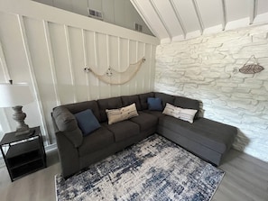 A sectional sofa with multiple cushions, a patterned rug, a small table with a lamp, and a hammock against a white wall with a stone texture.