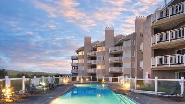 A swimming pool surrounded by lounge chairs, with a multi-story building in the background. - WM BLOCK Mariners Village 1br (Ocean Shores)