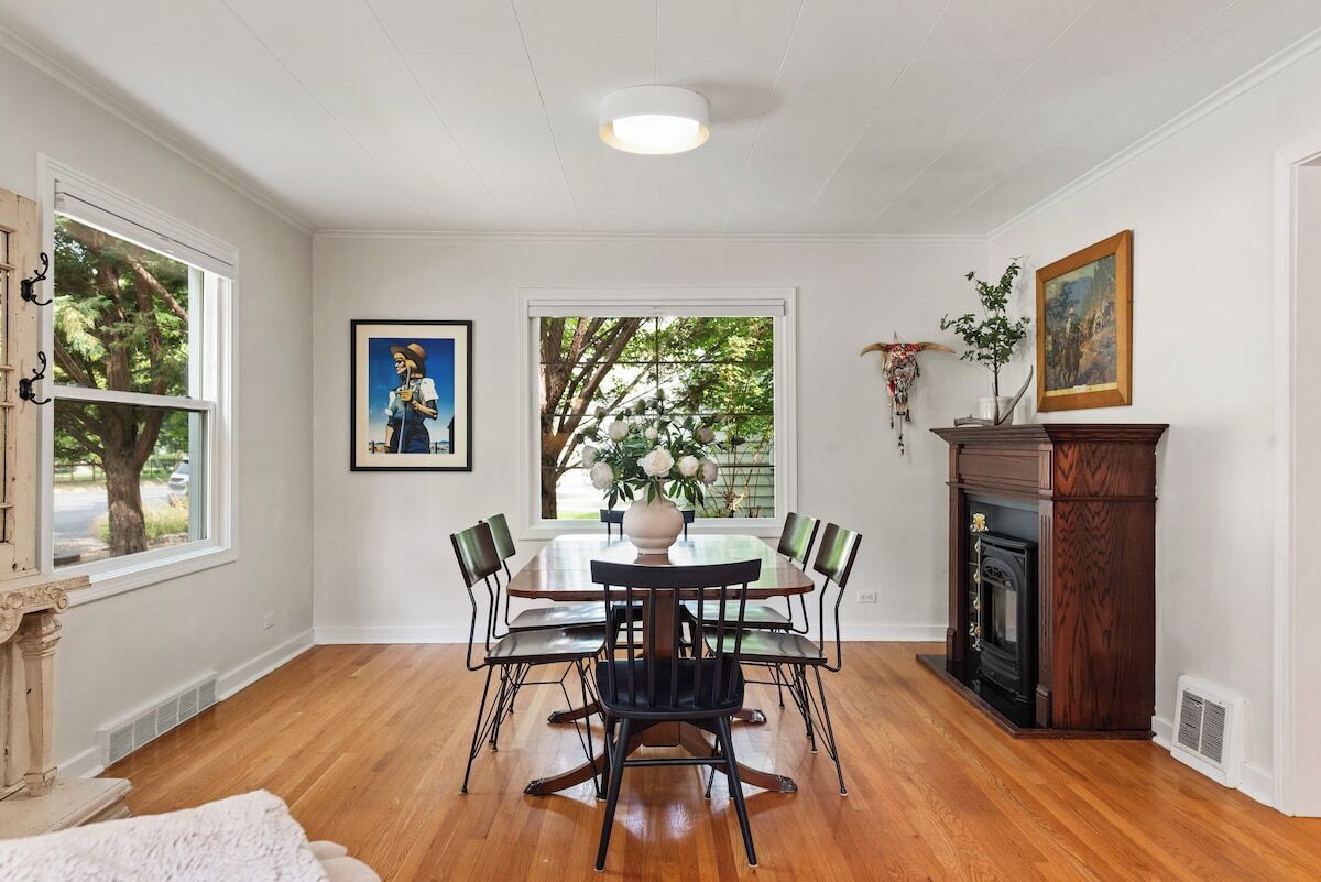 A dining room with a wooden table and chairs, a fireplace, and a window with a view of trees.