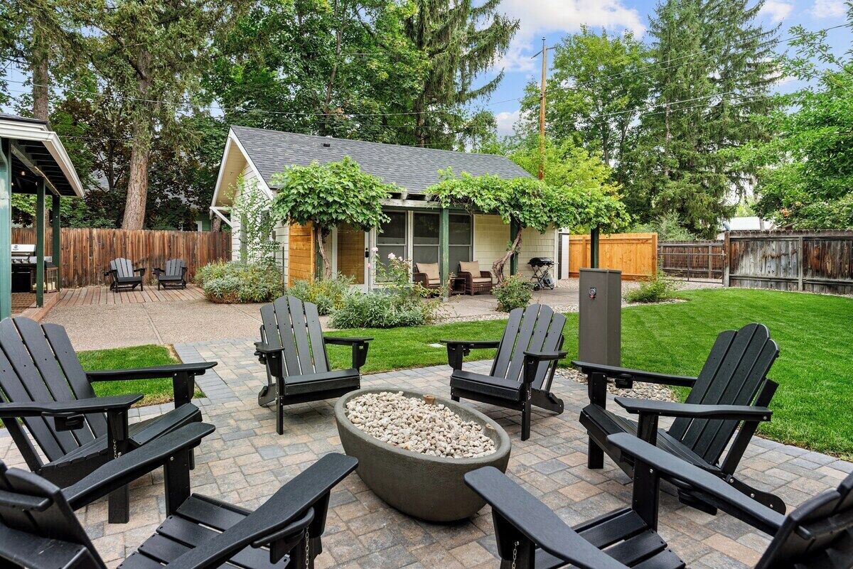 Un patio con sillas Adirondack, un brasero y una zona de estar cubierta con vistas a una casa y una valla de madera.