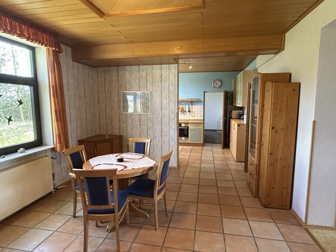 A dining area with a round table and chairs, a window with curtains, and a view of the kitchen in the background.