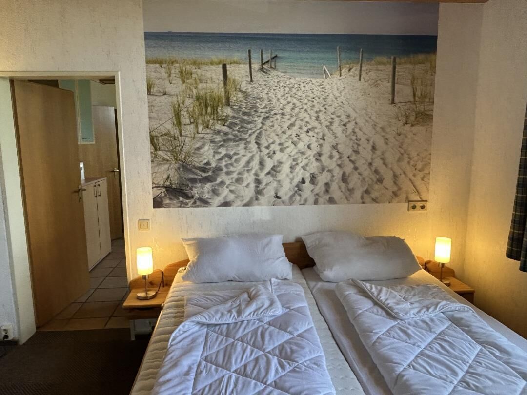 Ein Hotelzimmer mit einem großen Wandbild zum Strandthema, zwei Betten mit weißer Bettwäsche und Nachttischlampen.