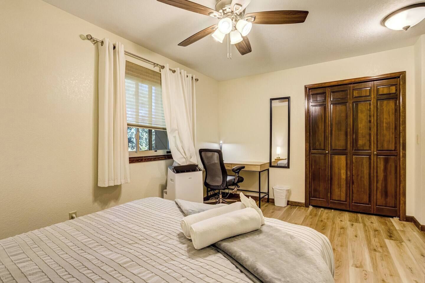 A hotel room with a bed, a desk, a chair, a ceiling fan, a window with curtains, and a wooden wardrobe.
