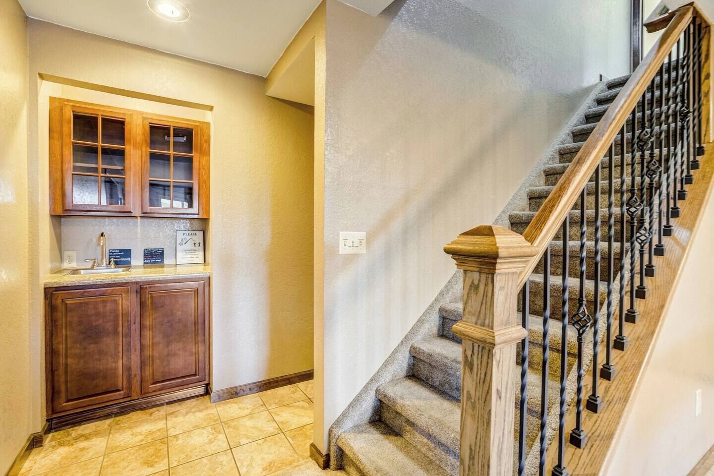 A kitchen with wooden cabinets and a countertop, a staircase with a wooden handrail and metal balusters, and a tiled floor.