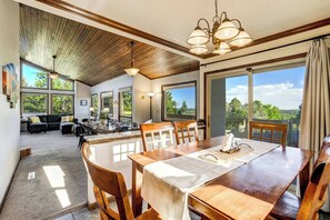 A spacious dining area with a wooden table and chairs, large windows offering a view of the outdoors, and a ceiling with wooden beams. - 6BR Sleeps 14 · Two Living Areas, Game Room, Hot Tub, Mtn View (Colorado Springs)