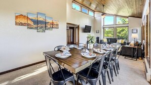 Uma sala de jantar com uma mesa comprida posta para várias pessoas, um sofá e uma televisão fixada na parede.