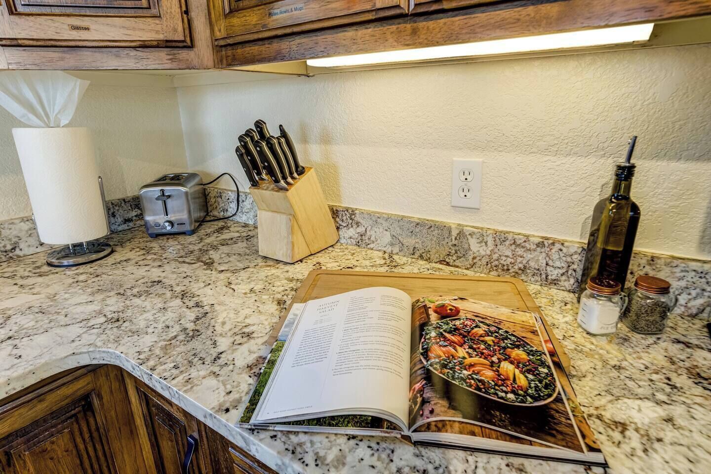 A kitchen with a granite countertop, a toaster, a knife block, a book, a baking tray with vegetables, and a bottle of olive oil.