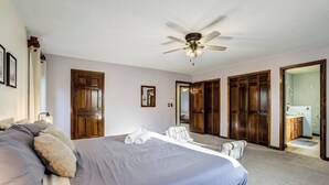 Quarto com uma cama grande, ventilador de teto, armários de madeira e banheiro visível por uma porta aberta.