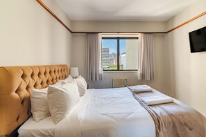 Une chambre d’hôtel avec un grand lit, une tête de lit, une fenêtre avec des rideaux, un téléviseur à écran plat fixé au mur et une vue sur les bâtiments environnants.