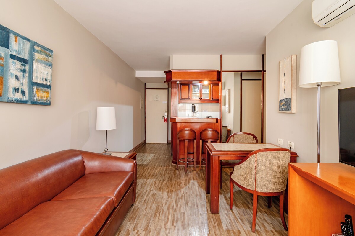 A hotel room with a leather sofa, a dining table with chairs, a kitchen area, and a flat-screen TV.