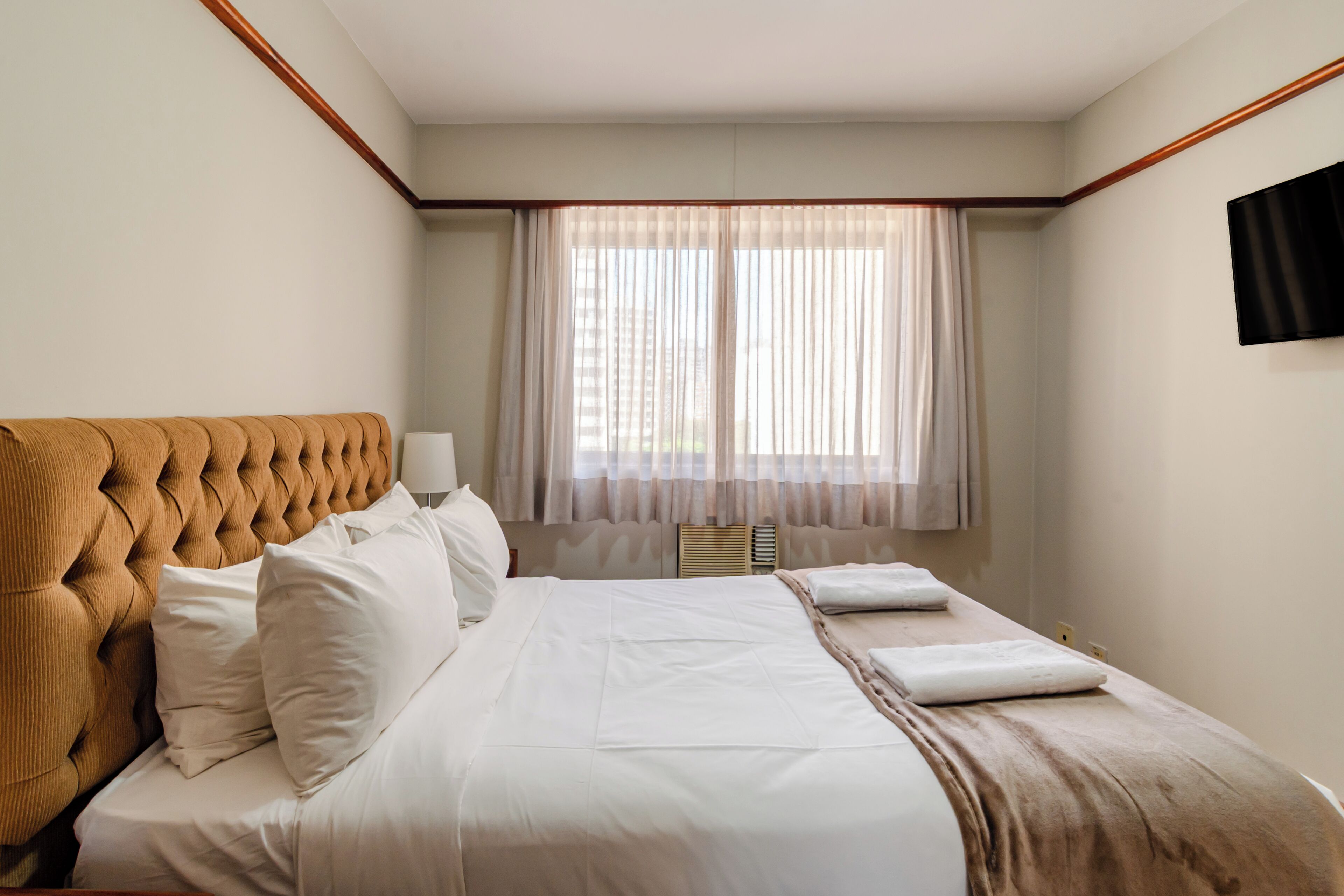 Une chambre d’hôtel avec un grand lit, une tête de lit, une fenêtre avec des rideaux légers et un téléviseur à écran plat fixé au mur.