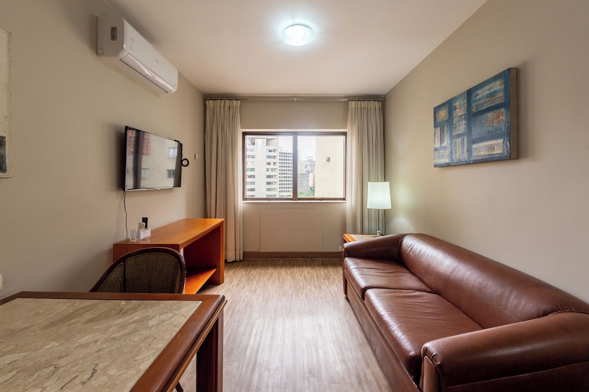 A hotel room with a wooden desk, a leather sofa, a television, a window with curtains, and a painting on the wall.