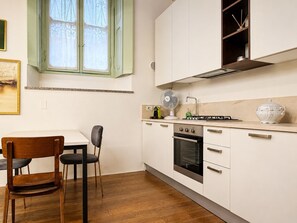 A modern kitchen with a gas stove, oven, and white cabinets. There is a dining table with chairs and a window with lace curtains.