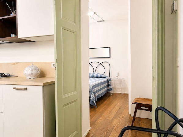 A narrow hallway leading to a bedroom with a bed and a bedside table. A kitchen area with a stove and a white pot is visible on the left.