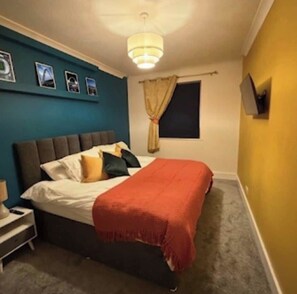 Ein Schlafzimmer mit Bett, Nachttisch, an der Wand montiertem Fernseher und einem Fenster mit Vorhängen.