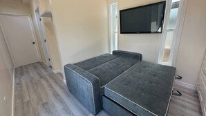 Habitación compacta con sofá cama gris, televisor de pantalla plana montado en la pared y suelo de madera.