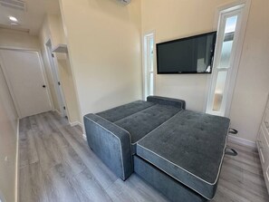 Habitación compacta con sofá cama gris, televisor de pantalla plana montado en la pared y suelo de madera.