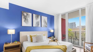 A hotel room with a large bed, two bedside tables, a nightstand, a balcony with a view of buildings, and three framed leaf artworks on the wall. - 2BR Intracoastal Retreat - Private Balcony (Hallandale Beach)