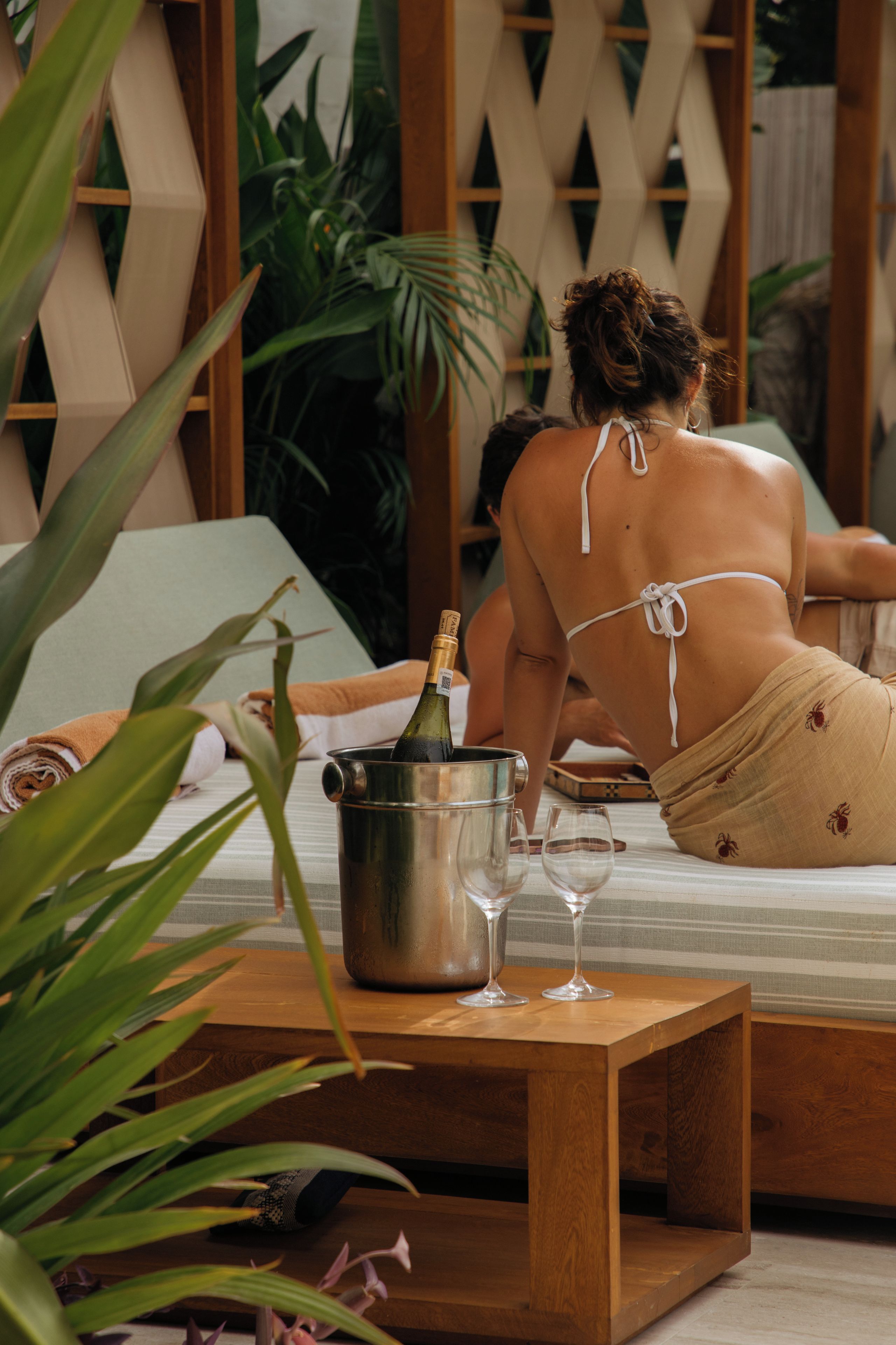 A person reclining on a bed with a bottle of champagne and two glasses on a wooden table.