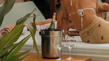 A person reclining on a bed with a bottle of champagne and two glasses on a wooden table.