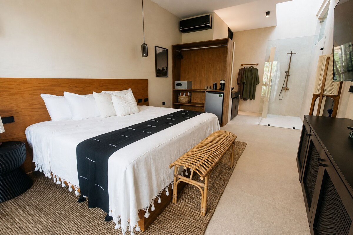 Quarto de hotel com cama, um banco, guarda-roupa, escrivaninha e área de chuveiro.