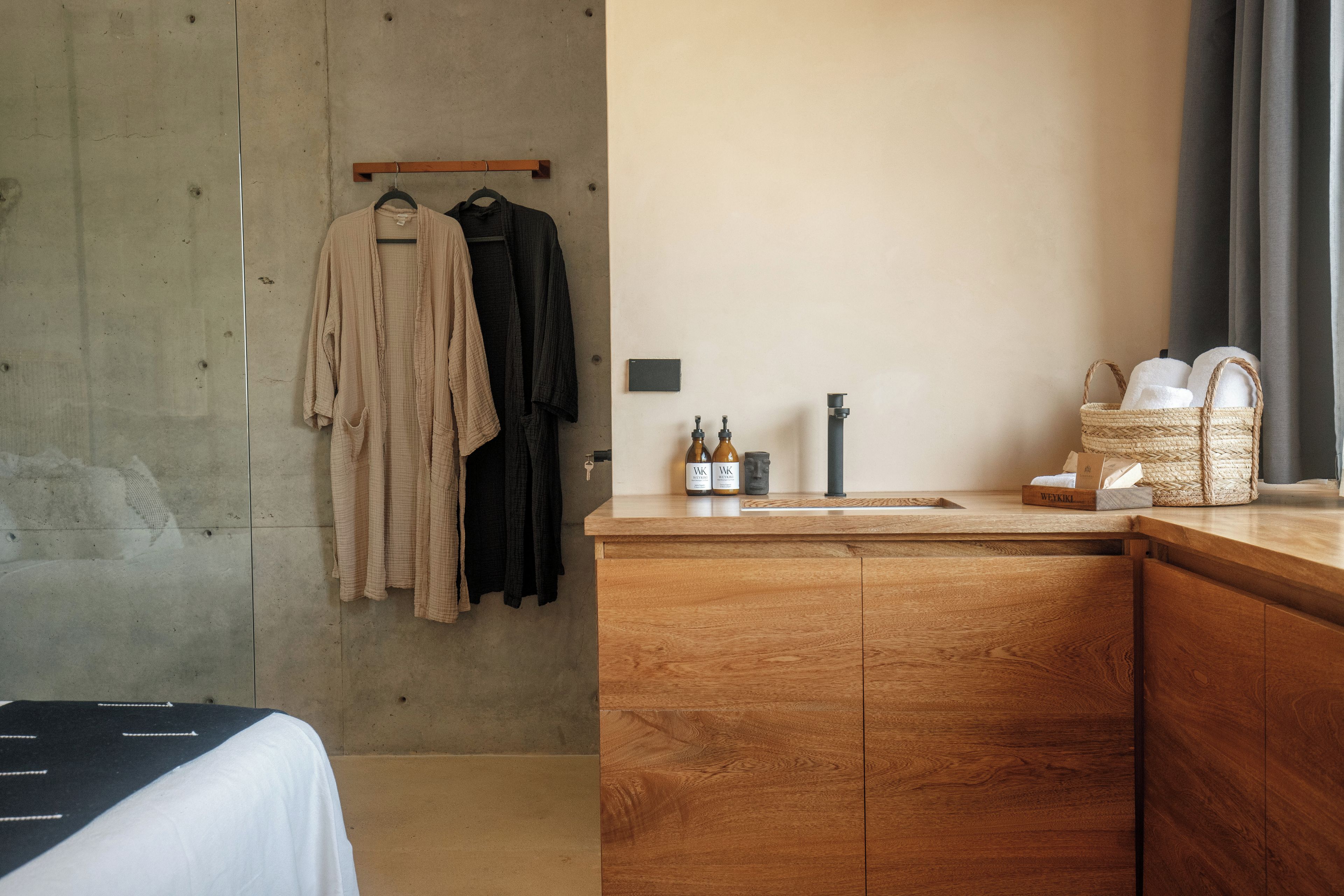 A hotel room with a wooden cabinet, a hanging rack with clothes, a small sink with bottles, and a window with curtains.