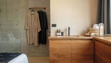 A hotel room with a wooden cabinet, a hanging rack with clothes, a small sink with bottles, and a window with curtains.