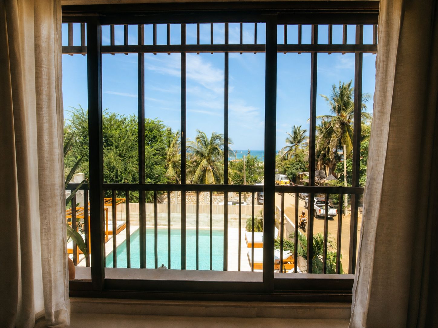 A window with metal bars, a pool, and a view of palm trees and a beach.