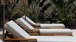 Fila de sillones de madera con cojines a rayas, dispuestos al aire libre frente a una pared de hormigón y plantas tropicales.