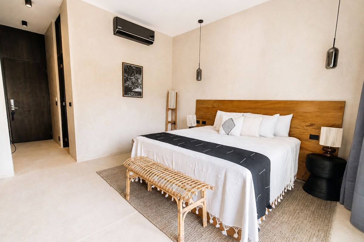 Un dormitorio con cabecera de madera, una cama con ropa de cama blanca, un banco, una lámpara de pie y un aire acondicionado mural.